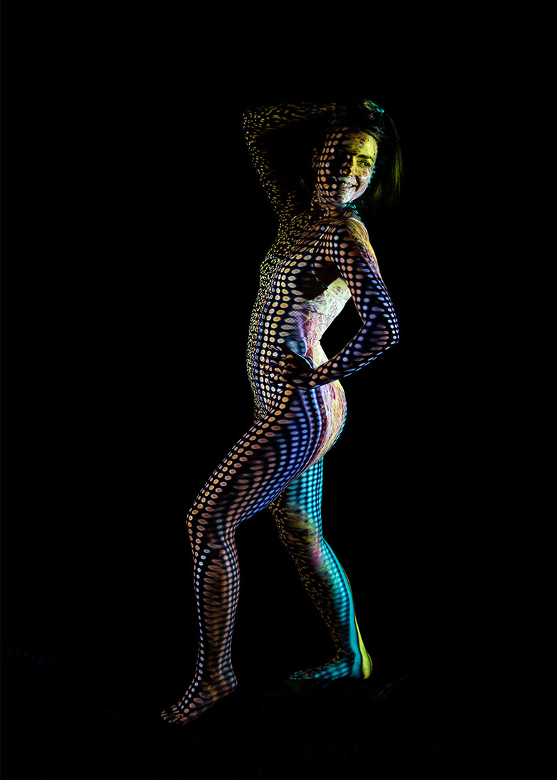 Person posing with one hand behind head and the other on the hip, covered in colorful dotted light patterns against a black background